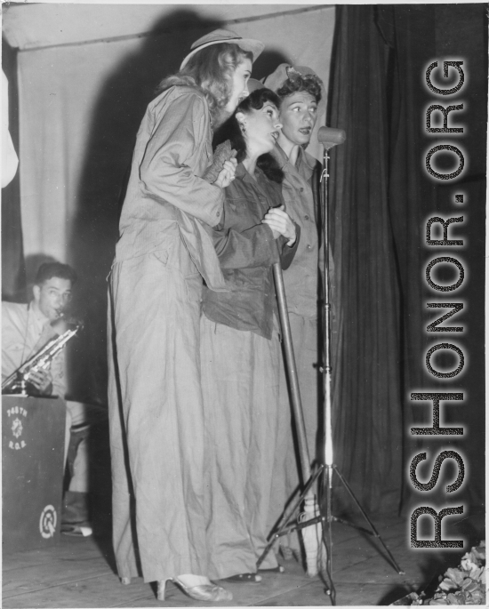 Ladies sing during USO performance in the CBI during WWII. GI plays sax by stage prop labeled for the 748th Railway Operating Battalion.