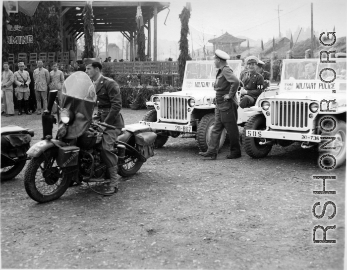 Burma Road dedication parade and ceremony in Kunming, China, on or around February 4, 1945, during WWII. Review of first convoy (or one of the first convoys) to reach China. General view of the stage area and MPs at the ready at jeeps provided by Service of Supply (SOS) .  The "Burma Road" was also often known as the "Stilwell Road," as it is in Chinese on the back of the stage, written “Shediwei Gonglu” (史迪威公路).