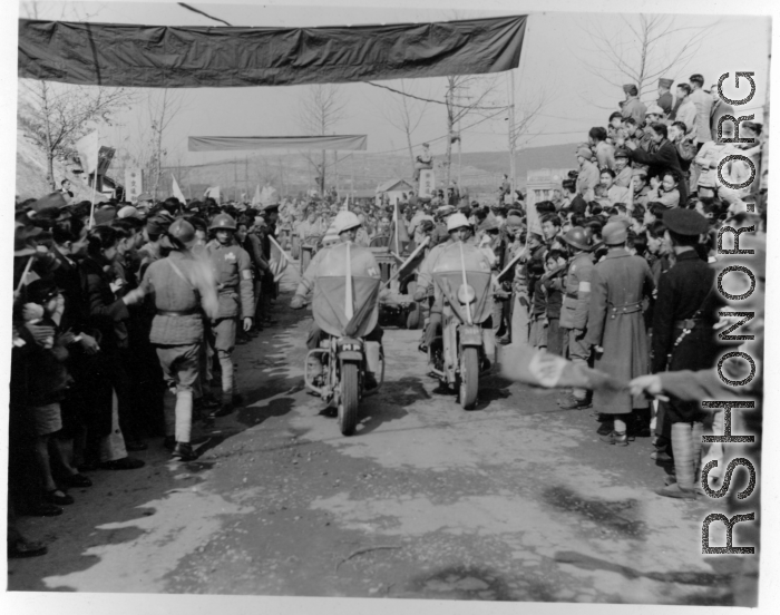 Burma Road dedication parade and ceremony in Kunming, China, on or around February 4, 1945, during WWII. Review of first convoy (or one of the first convoys) to reach China. Ranks of soldiers and civilians, waiting in anticipation of the parade, speeches, and first trucks to arrive, watch motorcycle MPs and jeeps, with US and Chinese flags.