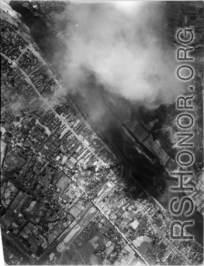 Bombing of small riverside town likely in SW China (esp. Guangxi), but possibly in Burma, or French Indochina. During WWII. Signs of previous bombings are visible about, including destroyed houses along the main road, and bomb craters here and there. Smoke arises in this image from recent bomb blasts.