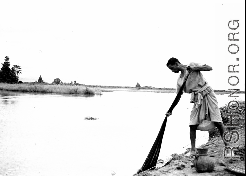 A man fishes with a next from the bank of a river. Probably in India. During WWII.