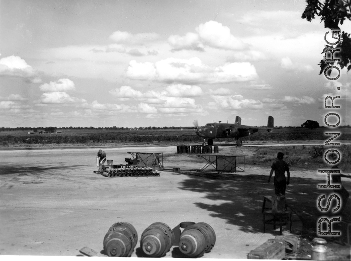 A B-25D of the American 491st Bombardment Squadron is on the taxiway.  Maintenance men who prepared it for the mission stand in the shade of tree. Small bombs, 100 pounds, are seen neatly stacked in the middle of the photo. Closer, in the center, are three 500 pound bombs. They lack the guide fins, and still have the transport 'rings' attached to make it easier to move them around.  Photo believed to have been taken at Yangkai, Yunnan Province, in the spring or summer of 1944. 