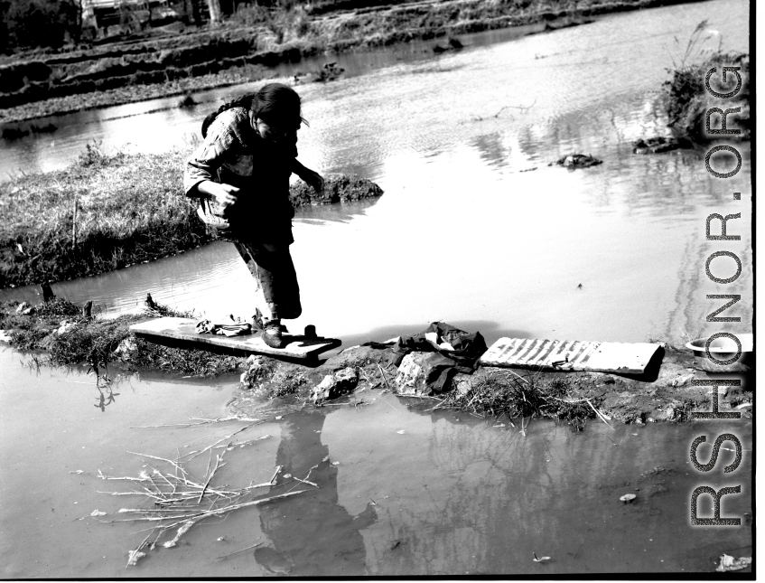 Local people in China: Elderly woman with bound feet crosses a precarious pathway of mud, boards, and stones. During WWII.  From the collection of Eugene T. Wozniak.