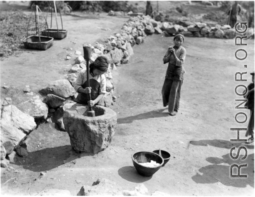 Local children laboring in China, Yunnan province, with a large mortar and pestle, either husking dry grain or mashing cooked rice so something similar. During WWII.