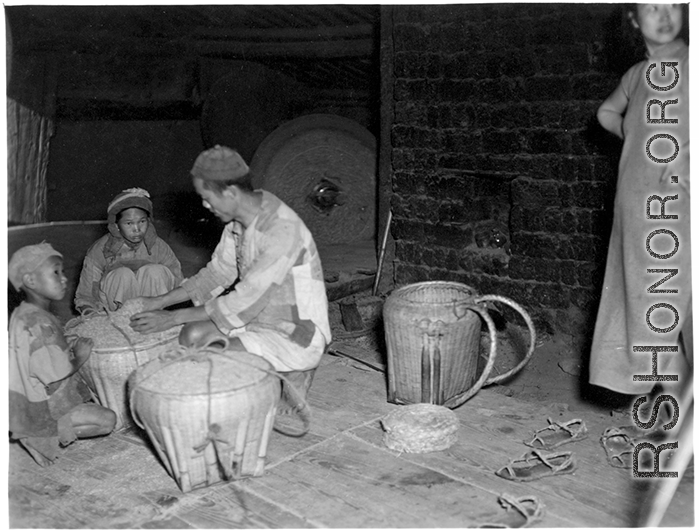 A family kneeling and working at a grinding mill in China during WWII, while a customer stands waiting. Note the large grinding wheel in motion behind them.