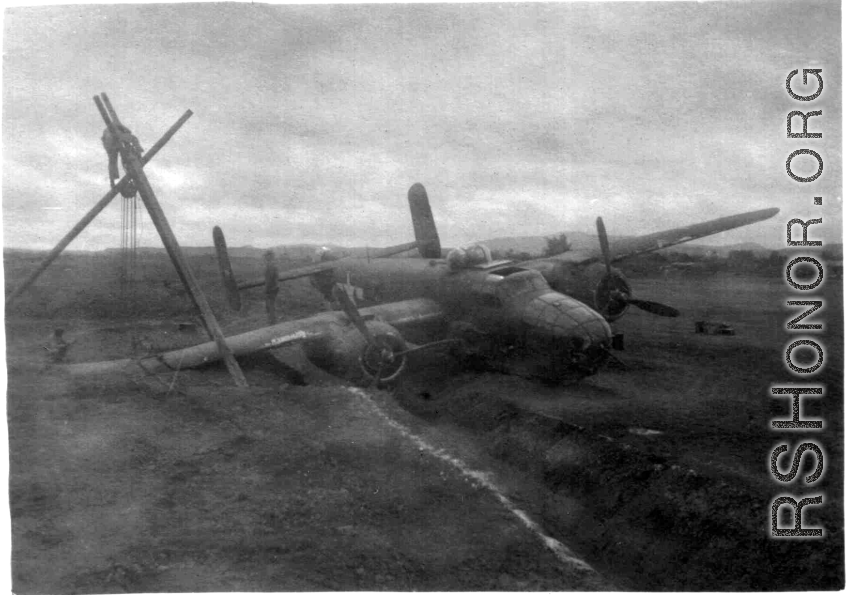 A B-25 in the ditch next to a runway or taxiway at an American base in China, during WWII. Crews work to salvage the aircraft using a simple human-powered tripod crane.