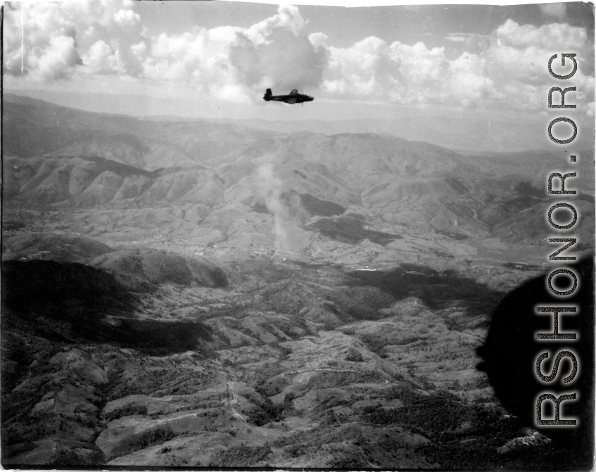 B-25 Mitchell bomber in flight in the CBI, in the area of SW China, Indochina, or Burma. Notice smoke rising in the center. During WWII.