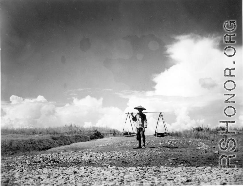 A farmer or laborer in China posing with a should pole. During WWII. Notice the freshly spread stone in the foreground, almost certainly crushed by hand, then hauled to the location by hand (for example, using shoulder poles), then spread and tamped down by human labor.