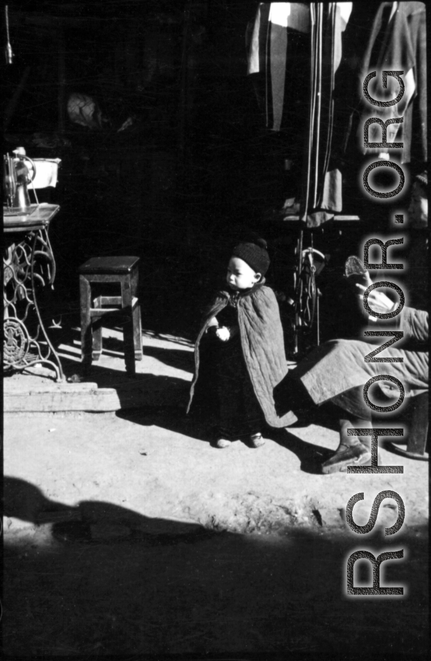 A young boy stands in the doorway of a seamstress shop in China, dressed in a warm cape. During WWII.