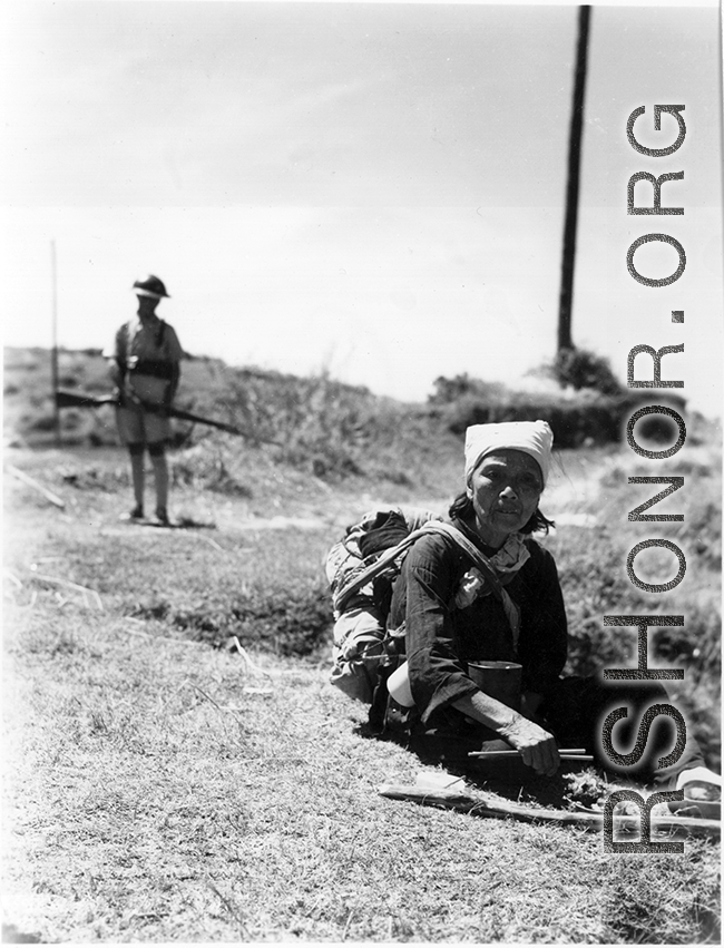 An elderly Chinese woman takes her lunch somewhere outside of Kunming, while a Nationalist guard is in the background. During WWII.