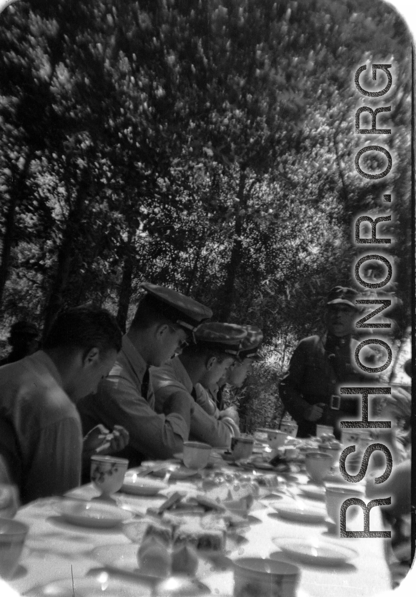 A Chinese officer makes a toast or speech as SACO members share a meal with Nationalist soldiers, in the shade of trees, during WWII. In northern China.