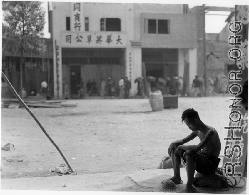 A tired looking young man sits in the opening to a stall advertising car parts, in Kunming, China, during WWII.