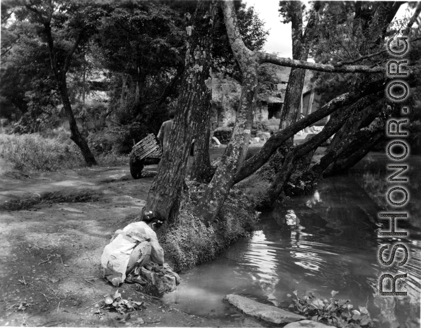 Local people in a village in Yunnan province, China: A woman washes clothes in a tree-shaded village pond. During WWII.