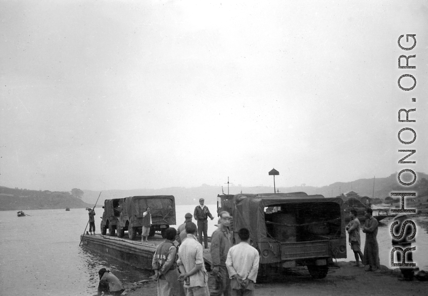 "Vehicles bound for Chengdu and the 20th bomber command are loaded on barges. As a steamboat (behind man directing) waits to ferry the barge across. All necessary gasoline was carried along for the journey. Several 55 gallon drums are visible in the vehicle. Poles were used to move the barges out of shallow water."