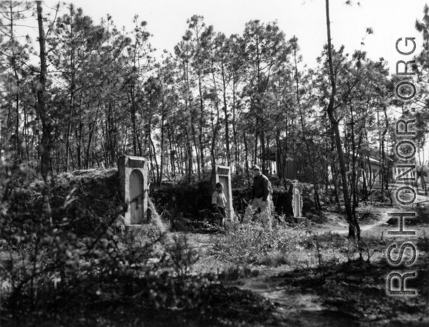 GI explorations of the hostel area at Yangkai air base during WWII: A GI walks past graves next to hostel buildings.