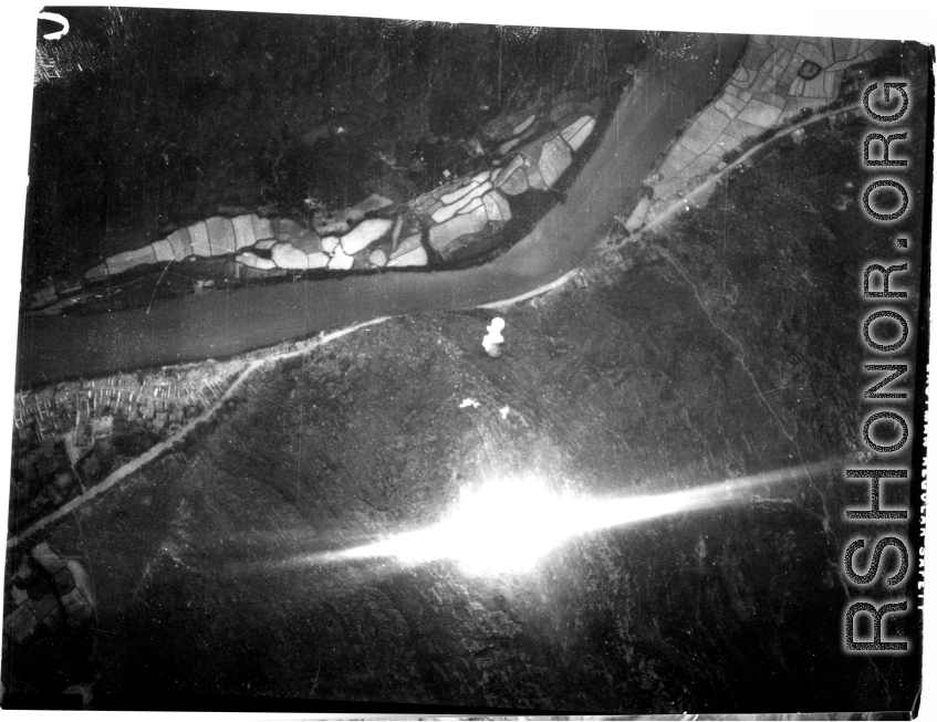 Bombing of small riverside town likely in SW China (esp. Guangxi), but possibly in Burma, or French Indochina. During WWII. Signs of previous bombings are visible about, including destroyed houses along the main road, and bomb craters here and there. The bright white spot is the arc flash of a bomb explosion, caught exactly at the right time, a another explosion is starting just above it.