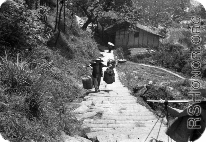 A man carries large packages up a steep mountain path using a shoulder pole in China during WWII.