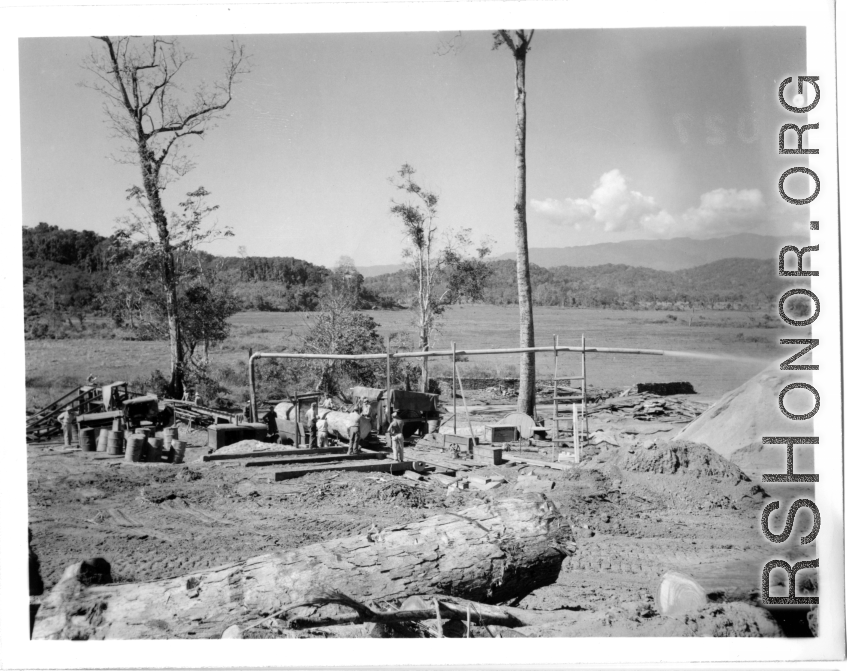 Site at the mill yard, including a large log heading towards saw at a lumber mill of the 797th Engineer Forestry Company in Burma. Sawdust flys through pipe onto large pile.  During WWII.