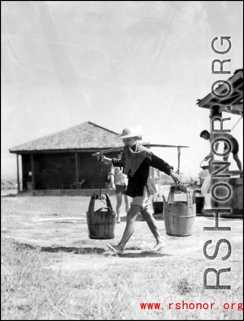 A Chinese staff person at an American base in China during WWII carries buckets on a shoulder pole.