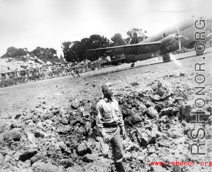 Kenneth Williams inspects a bomb crater at an American base in China during WWII. A C-46 transport is in the background.