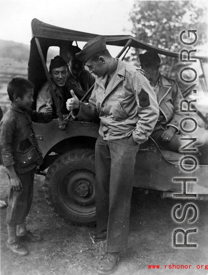 GI exchanges a thumbs up with kid in China, while other GIs observe from jeep. During WWII.