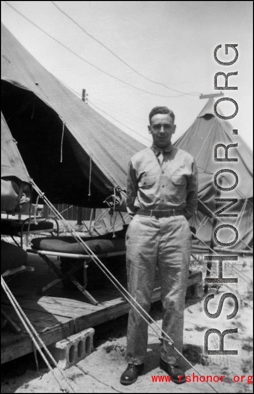 An American stands next to a tent with bed cots, in the CBI, during WWII.