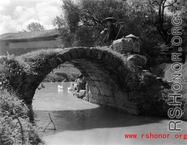 A rural woman shouldering baskets crosses a worn footbridge in China.  From the collection of Eugene T. Wozniak.