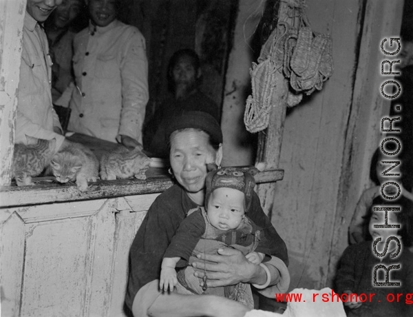 A woman holds a small child in a shop in Yunnan, China, during the Second World War.  Note the straw sandals hanging on the shop window in the back.