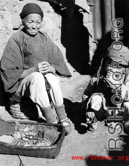 Local people in China: An elderly woman---possibly working as a petty seamstress doing minor repairs on shoes or cloth items---sits with child. During WWII.
