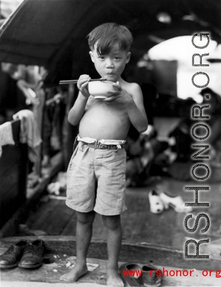 A young boy eats a meal on a boat in China during WWII, probably in Guangxi province, likely Liuzhou or Guilin.