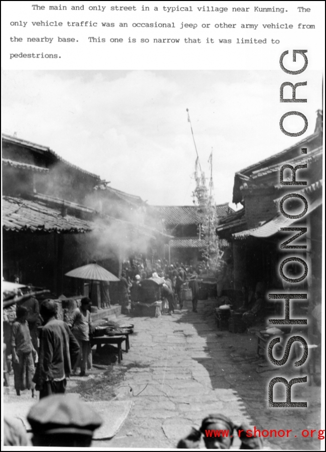 A very narrow main market street in a typical village near Kunming, Yunnan province. In addition to food stalls and other sellers, in this case a funeral activity (usually several days long) is being carried out in part in the street. During WWII.