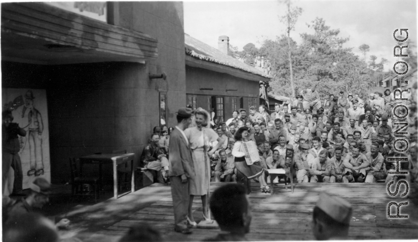 Celebrities (including Ann Sheridan, Ben Blue, and Ruth Dennis on instrument in this shot) perform on an outdoor stage set up at the "Last Resort" at Yangkai, Yunnan province, during WWII. Notice both Americans and Chinese in the audience for this USO event.