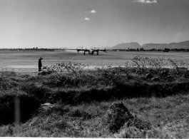 B-24 stirs up dust on runway at Kunming, China, during WWII. Chinese worker stands in the foreground, as well as some in the far distance.