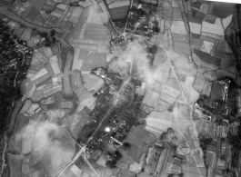 Bombs explode on a compact village in the midst of rice paddies in either SW China (esp. Guangxi), or Burma, or French Indochina during WWII. The bright white spot is the arc flash of a bomb explosion, caught exactly at the right time.