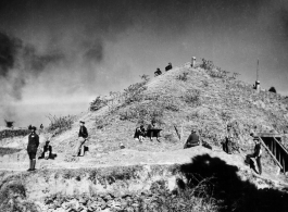 Pyramid-shaped air raid shelter used by Chennault in Kunming during WWII, in the midst of an air raid. In this image, Chennault is standing toward left, looking at the Japanese bombing out of scene of the left. In the middle, an American GI makes plots on a map board. Air vents can be seen protruding from the top, and garden vegetables grow in front.  Note also the female professional staff. A woman, probably a westerner, sits next to the GI with the map board, and another woman, likely of Chinese descent, 