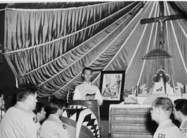 A parachute forms a backdrop in this Chapel in Kunming in 1945. Father Charles F. Meeus celebrates mass.  The Father's pulpit is the cowling for the P-40K "Little Flower" flown by Ray Kaiser (25th FS). "Little Flower" was St Therese from Lisieux, France. (Thanks JBarbaud)  Notice the corrugated metal making up the lower area of the alter, also likely parts from aircraft or packaging munitions.