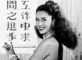 Jinx Falkenburg poses with a classroom slogan that says, roughly, "from cooperation seek progress in learning." In China during WWII.