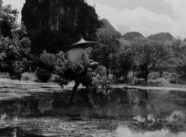 A Chinese man fishes from upon a narrow branch over a pond, with a karst peak in the background, in SW China during WWII.