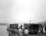 "Vehicles bound for Chengdu and the 20th bomber command are loaded on barges. As a steamboat (behind man directing) waits to ferry the barge across. All necessary gasoline was carried along for the journey. Several 55 gallon drums are visible in the vehicle. Poles were used to move the barges out of shallow water."