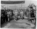 Burma Road dedication parade and ceremony in Kunming, China, on or around February 4, 1945, during WWII. Review of first convoy (or one of the first convoys) to reach China. Ranks of soldiers and civilians, waiting in anticipation of the parade, speeches, and first trucks to arrive, watch motorcycle MPs and jeeps, with US and Chinese flags.
