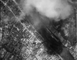 Bombing of small riverside town likely in SW China (esp. Guangxi), but possibly in Burma, or French Indochina. During WWII. Signs of previous bombings are visible about, including destroyed houses along the main road, and bomb craters here and there. Smoke arises in this image from recent bomb blasts.