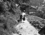 A man carries large packages up a steep mountain path using a shoulder pole in China during WWII.