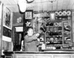 Man in the "Zhang family" hat shop does something to a hanging hat. During WWII. In SW China.