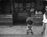 A child relieving itself streetside, in front of a shop, in China during WWII.