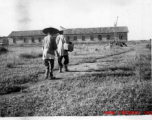 Two women with bound feet walking at an American base in Yunnan province, China, during WWII, with a barracks or other building in the background.