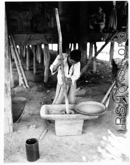 Local people in Burma near the 797th Engineer Forestry Company--A man is working most likely to dehusk grain, or to make some other worked produce, while another person waits with a woven dry pan.  During WWII.