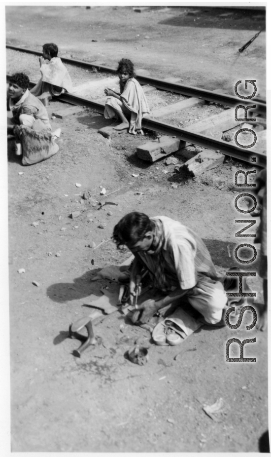 Man does shoe repair with simple tools next to the railway. In India.  Scenes in India witnessed by American GIs during WWII. For many Americans of that era, with their limited experience traveling, the everyday sights and sounds overseas were new, intriguing, and photo worthy.