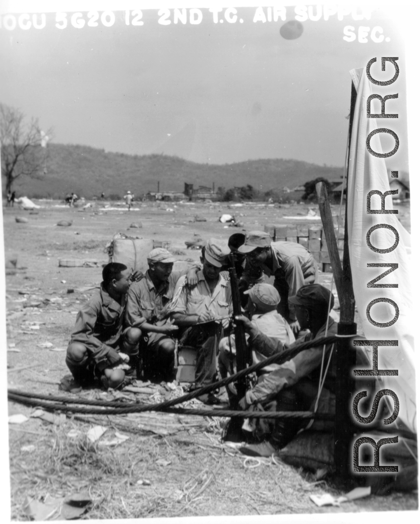 On the ground in the midst of air drop parachutes, a GI coordinates with a group of Chinese soldiers.