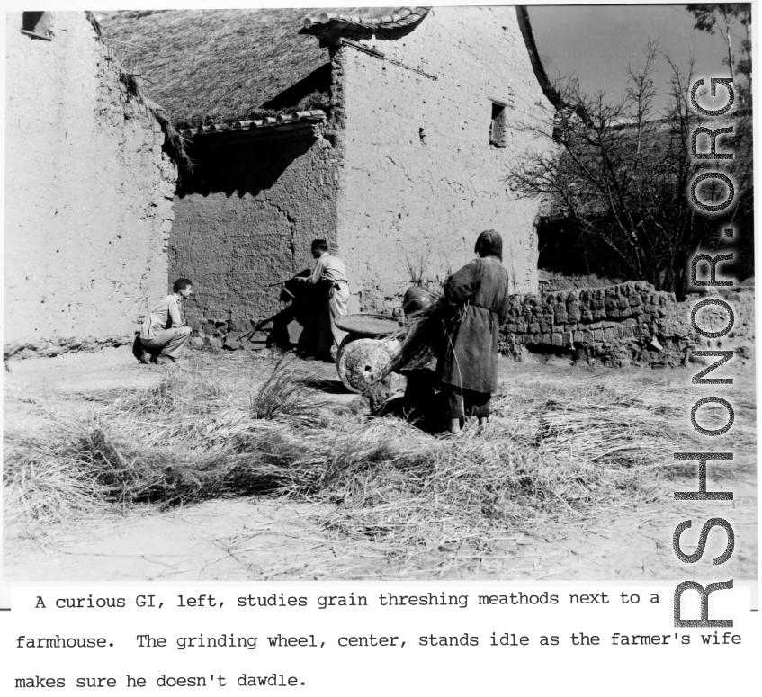 A GI points his carbine down the narrow space between two homes in a Chinese village during WWII, as a local village woman threshes grain.