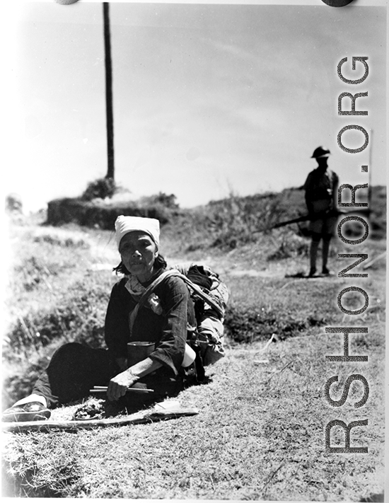 A woman in Yunnan province, China, rests to eat a meal, during the hard days of WWII.  Note the foot container on her lap and the chopsticks in her hands. A soldier with gun stands in the background.  From the collection of Eugene T. Wozniak.